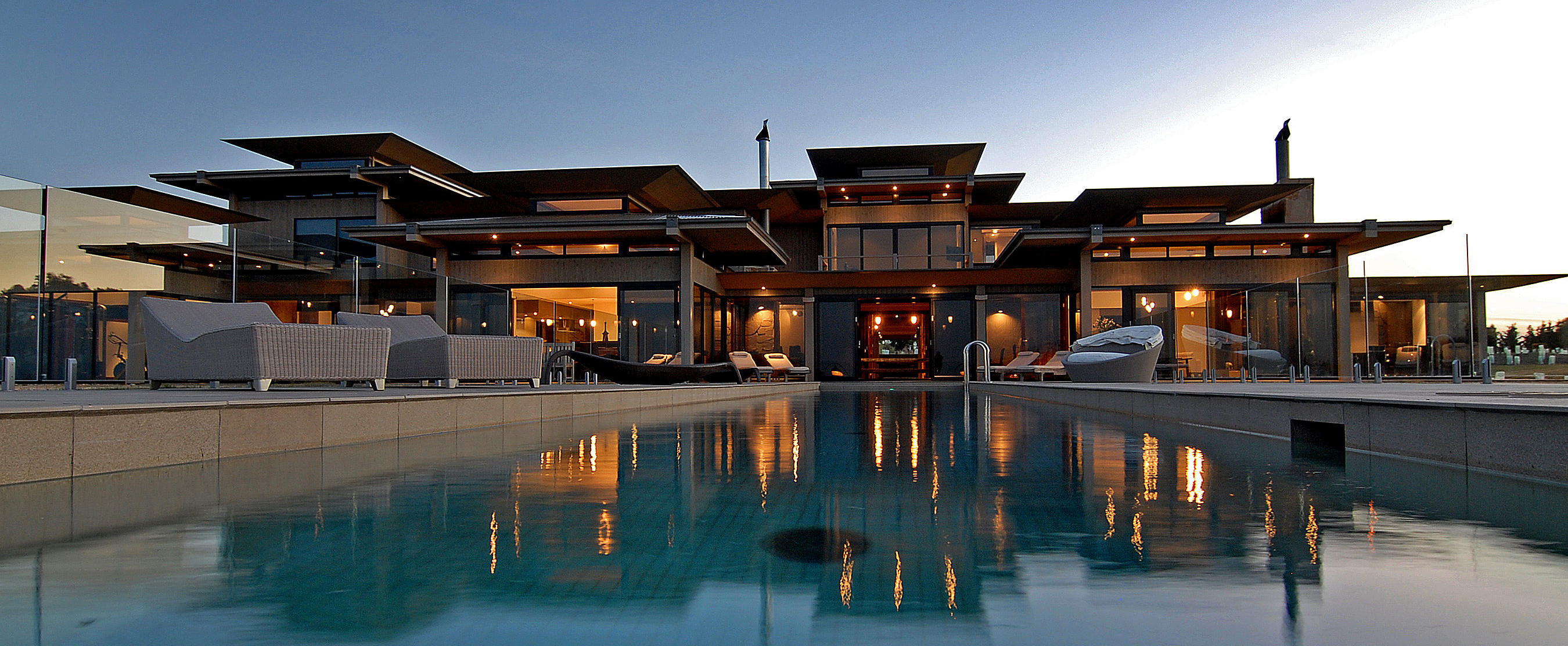 Villa privée élégante en Australie au coucher du soleil, avec piscine éclairée, grand patio meublé et architecture moderne en verre et bois.