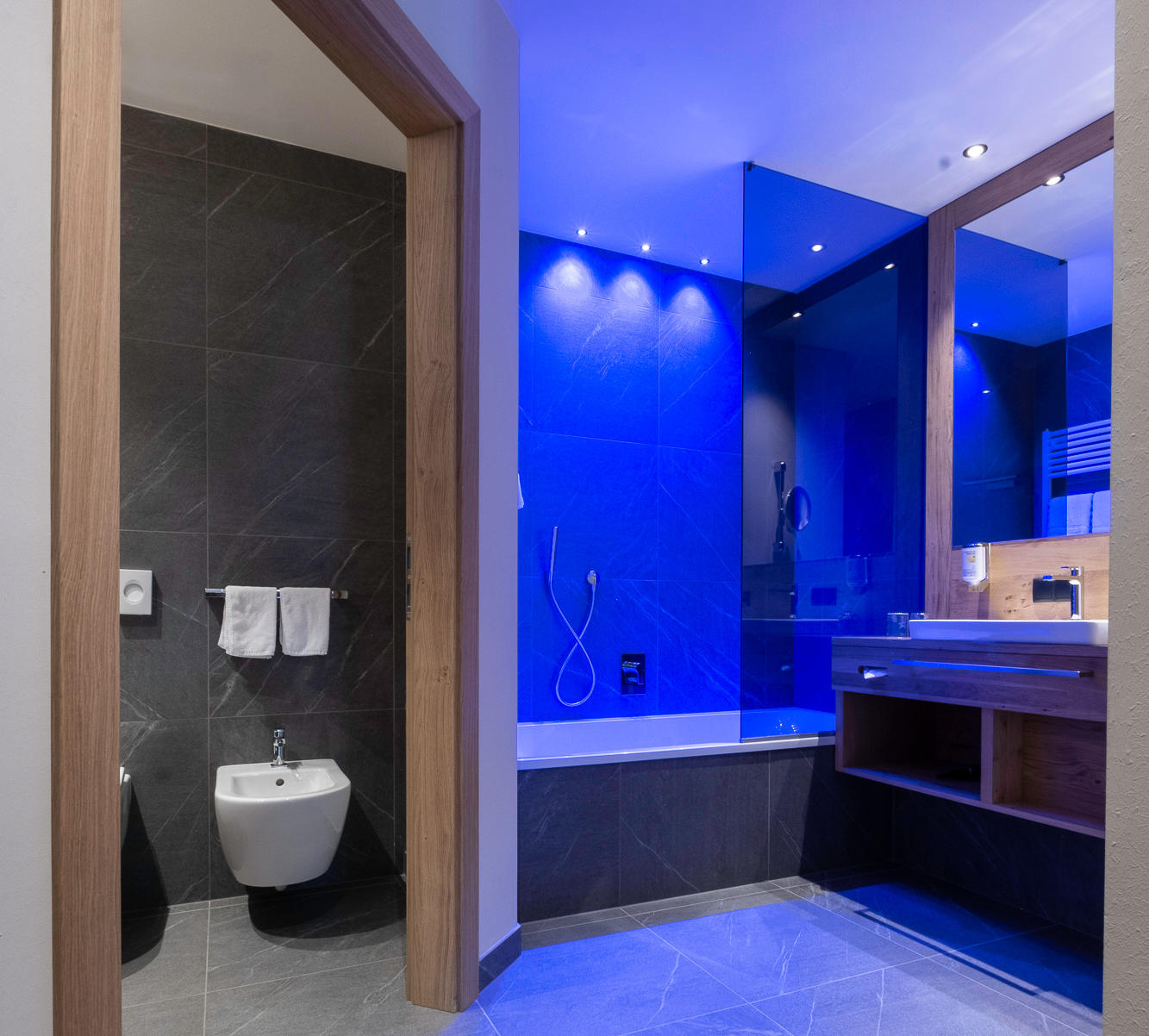 Elegant modern bathroom of the Hotel Terentnerhof with blue lighting, grey tiles, bathtub, bidet and sink.