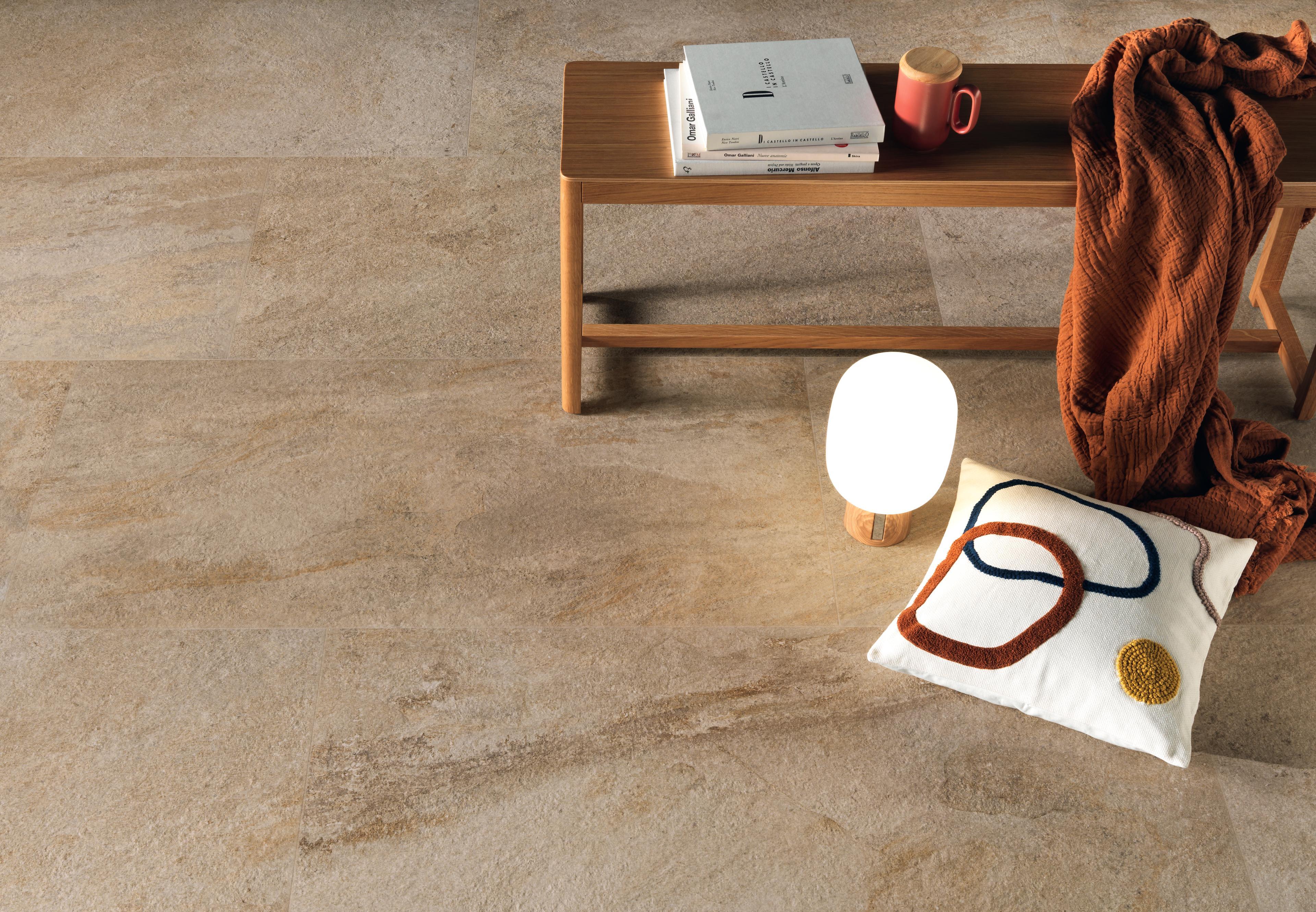 Floor with beige tiles with golden veins, furnished with a wooden table on which are resting books and a red cup. 