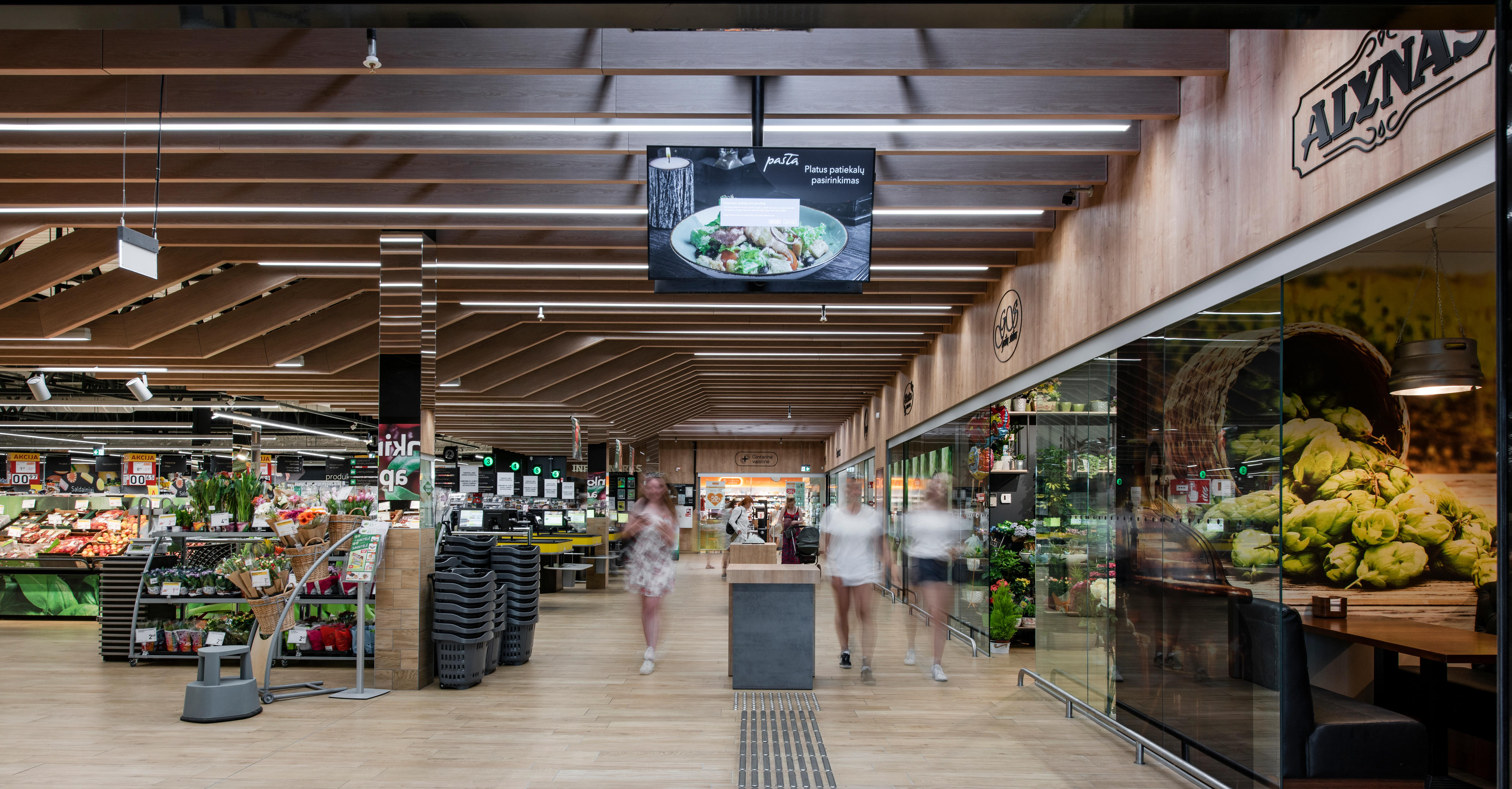 Interno di un supermercato con scaffali di prodotti freschi, carrelli della spesa impilati e clienti che camminano lungo il corridoio centrale.