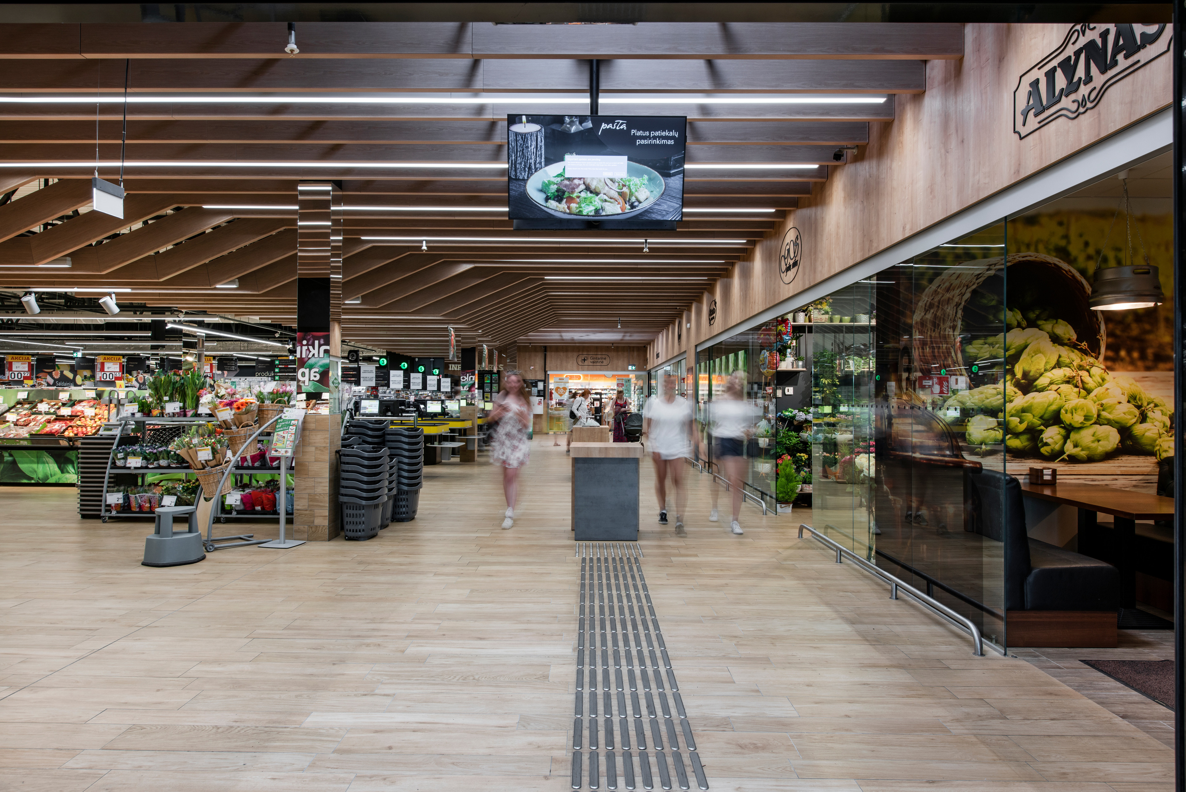 Интерьер супермаркета с полками, заполненными свежими продуктами, тележками и покупателями, идущими по проходу.