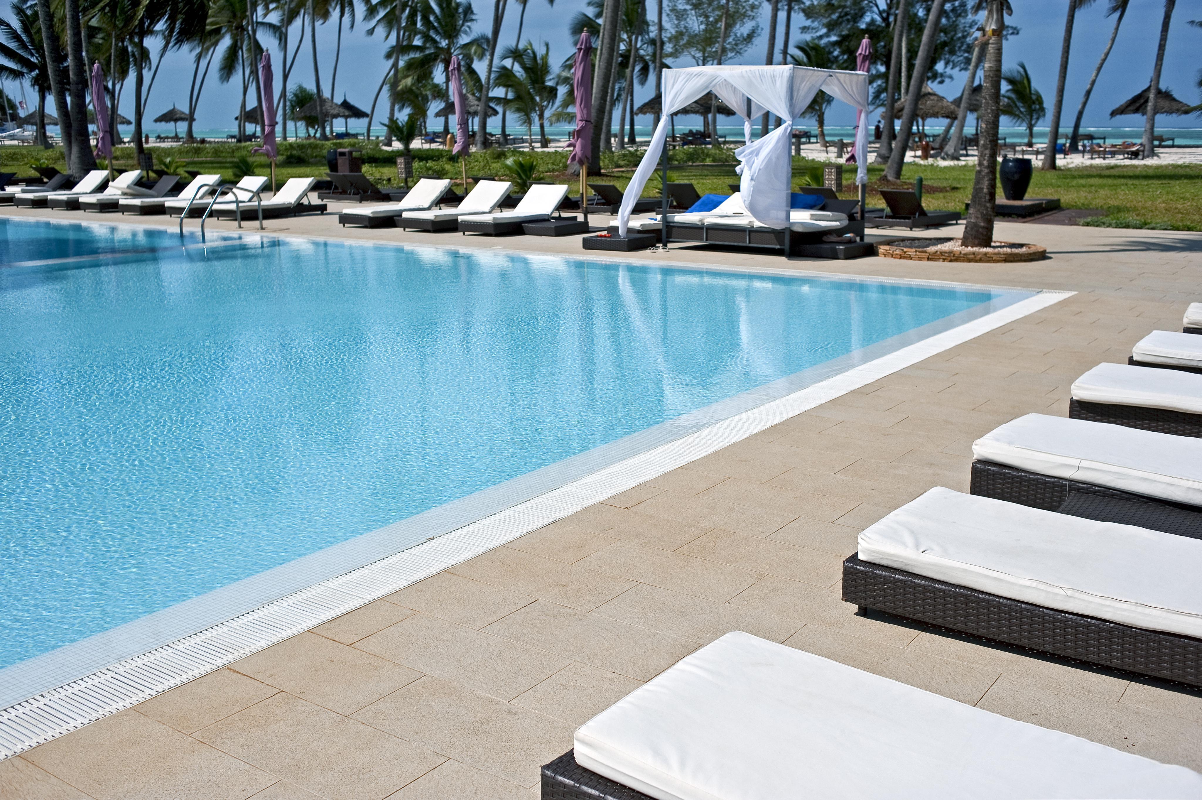 Outdoor pool at the Diamond Dream resort in Zanzibar with white sun loungers and a gazebo, surrounded by palm trees and overlooking the sea.