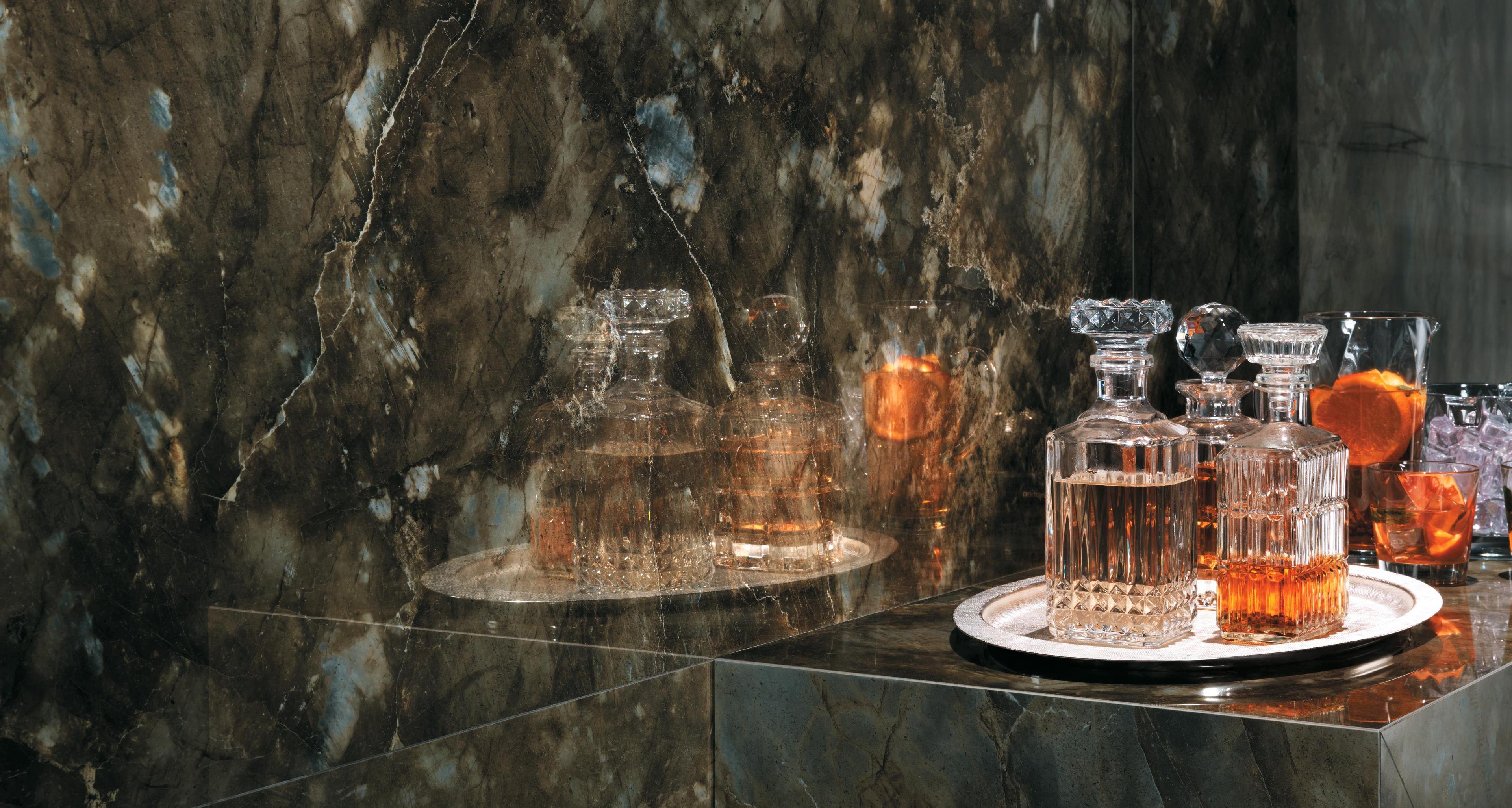 Pared de gres porcelánico efecto mármol verde con reflejos, decorada con bandejas de cristales y vasos de licor.