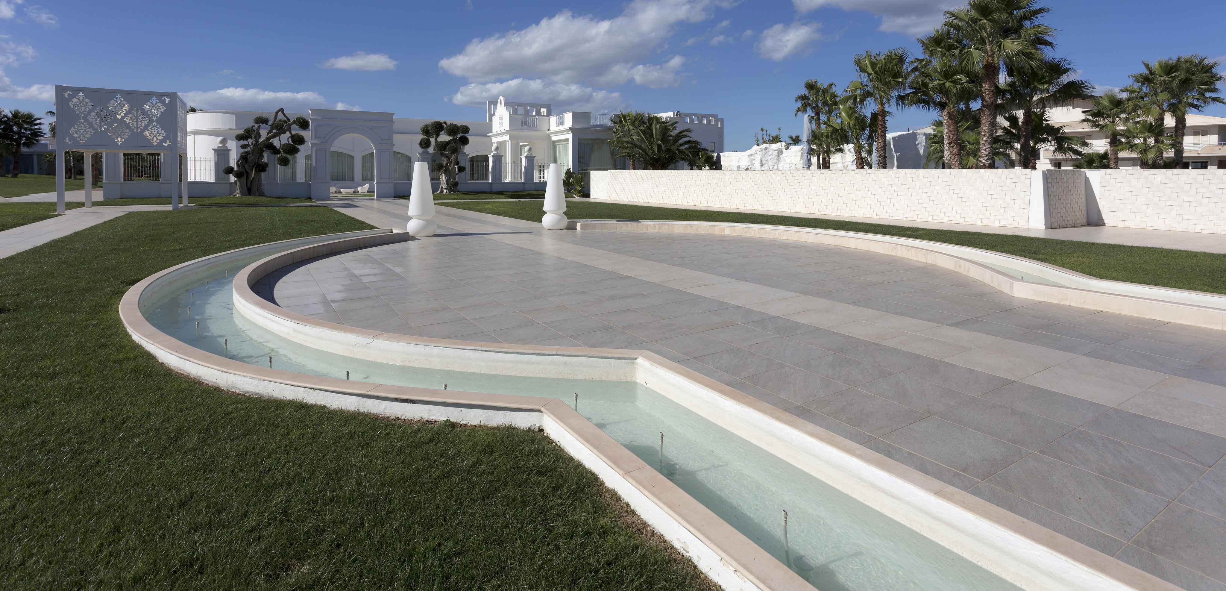  La cour du bâtiment Pietra Perla de Sannicandro di Bari, avec un jardin pavé, des sculptures blanches, des palmiers et un ciel bleu. 