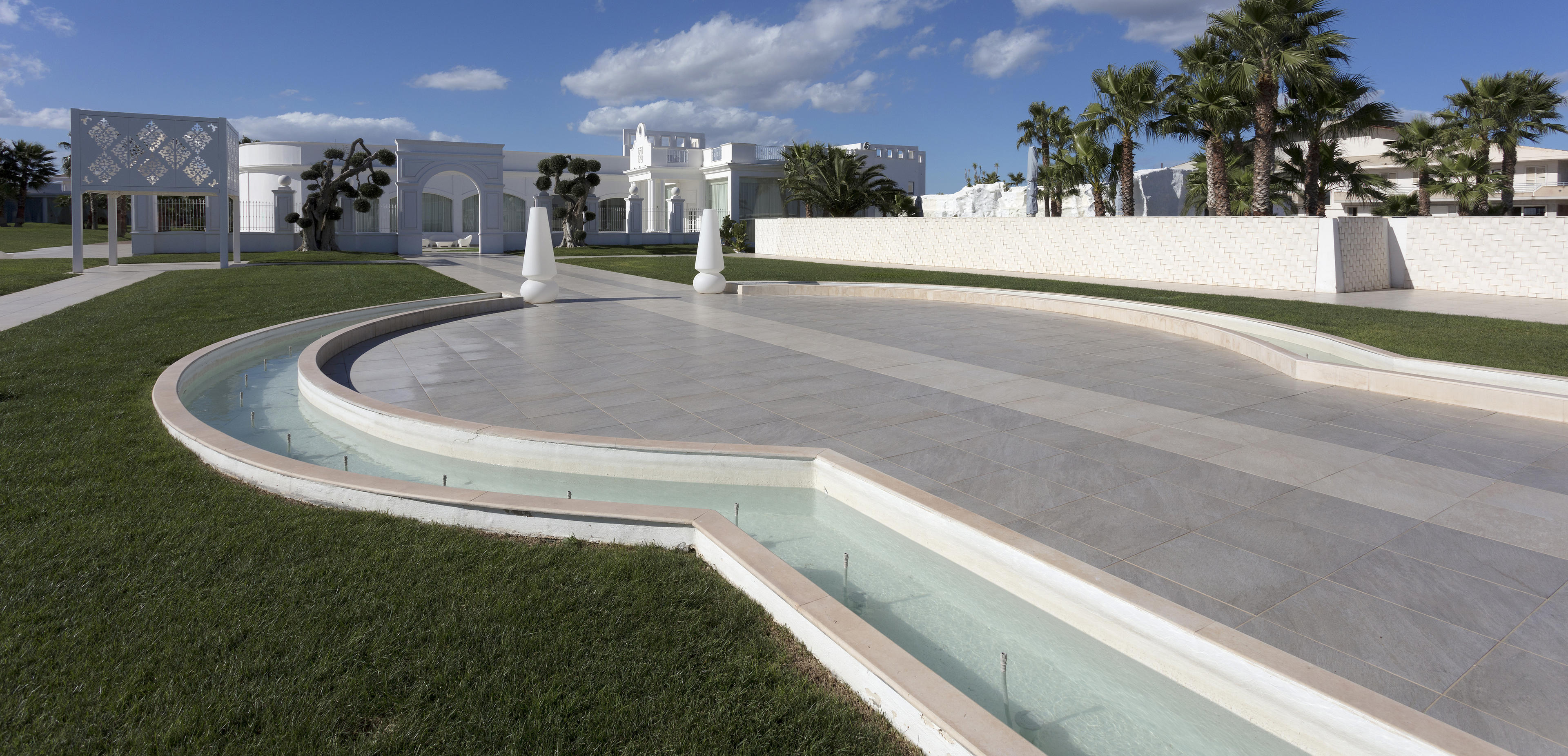La cour du bâtiment Pietra Perla de Sannicandro di Bari, avec un jardin pavé, des sculptures blanches, des palmiers et un ciel bleu.