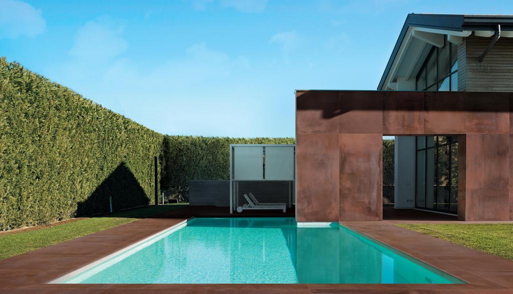 Piscina moderna con piastrelle antiscivolo color terracotta, circondata da siepi verdi e un'area relax con lettini e pergola bianca.