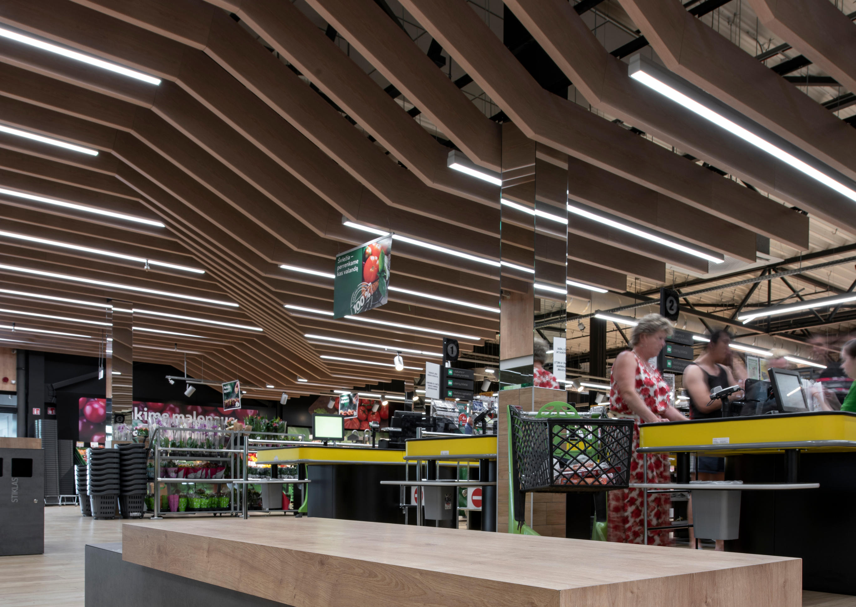 Interno di un supermercato con un banco di legno moderno in primo piano e clienti alle casse sullo sfondo.