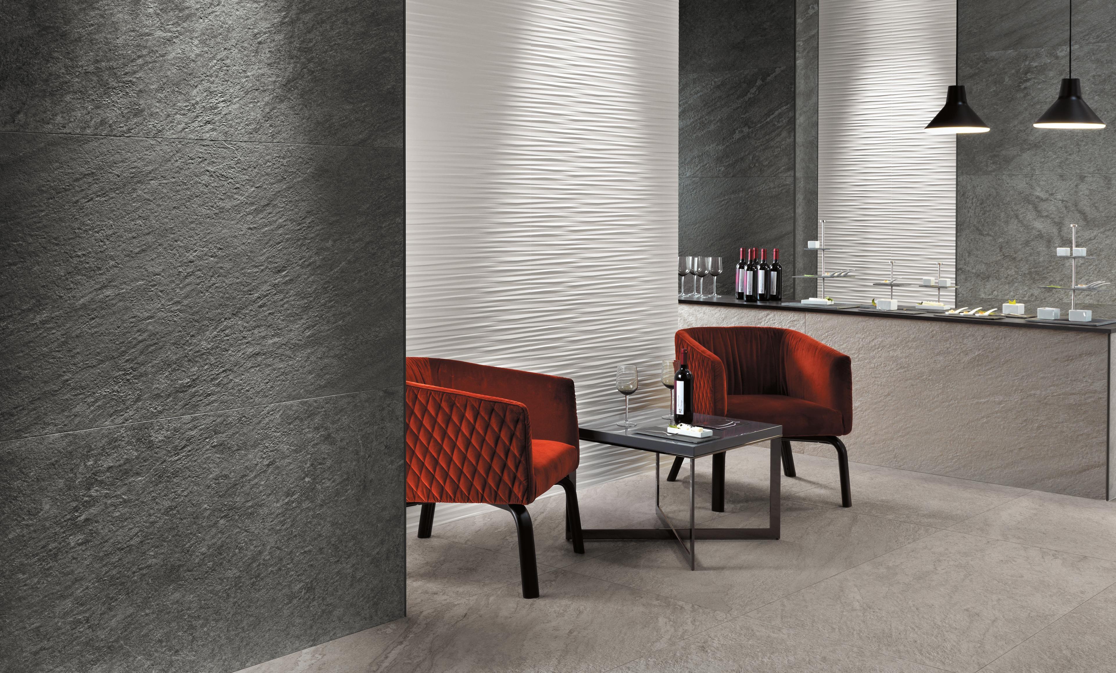 Intérieur élégant avec sol et murs en grès cérame Atlas Concorde Brave. Deux fauteuils rouges sont disposés autour d’une petite table, avec des bouteilles de vin sur le comptoir en arrière-plan.