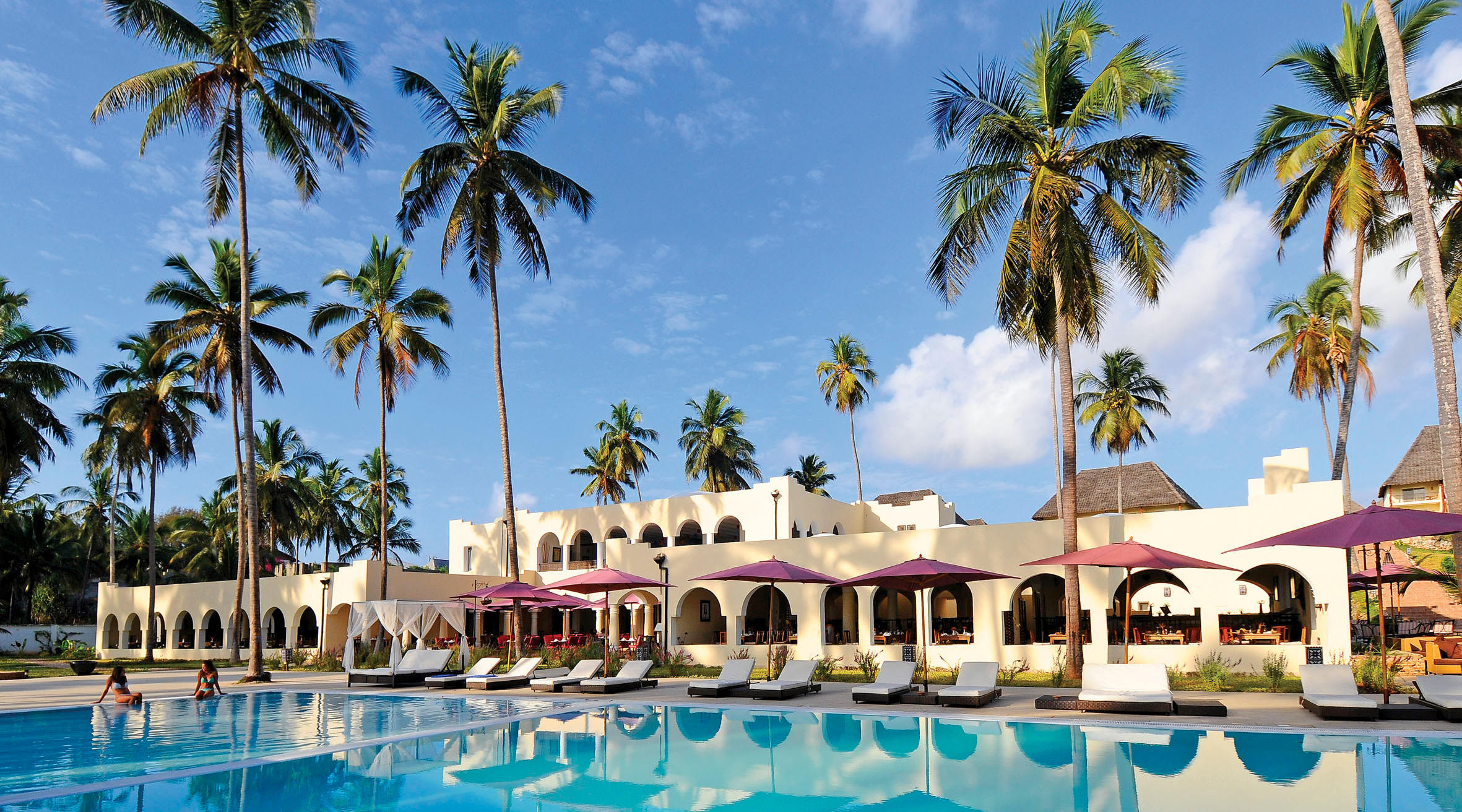 Outdoor pool at the Diamond Dream resort in Zanzibar, surrounded by palm trees and sun loungers, with buildings of Arabic architecture in the background.