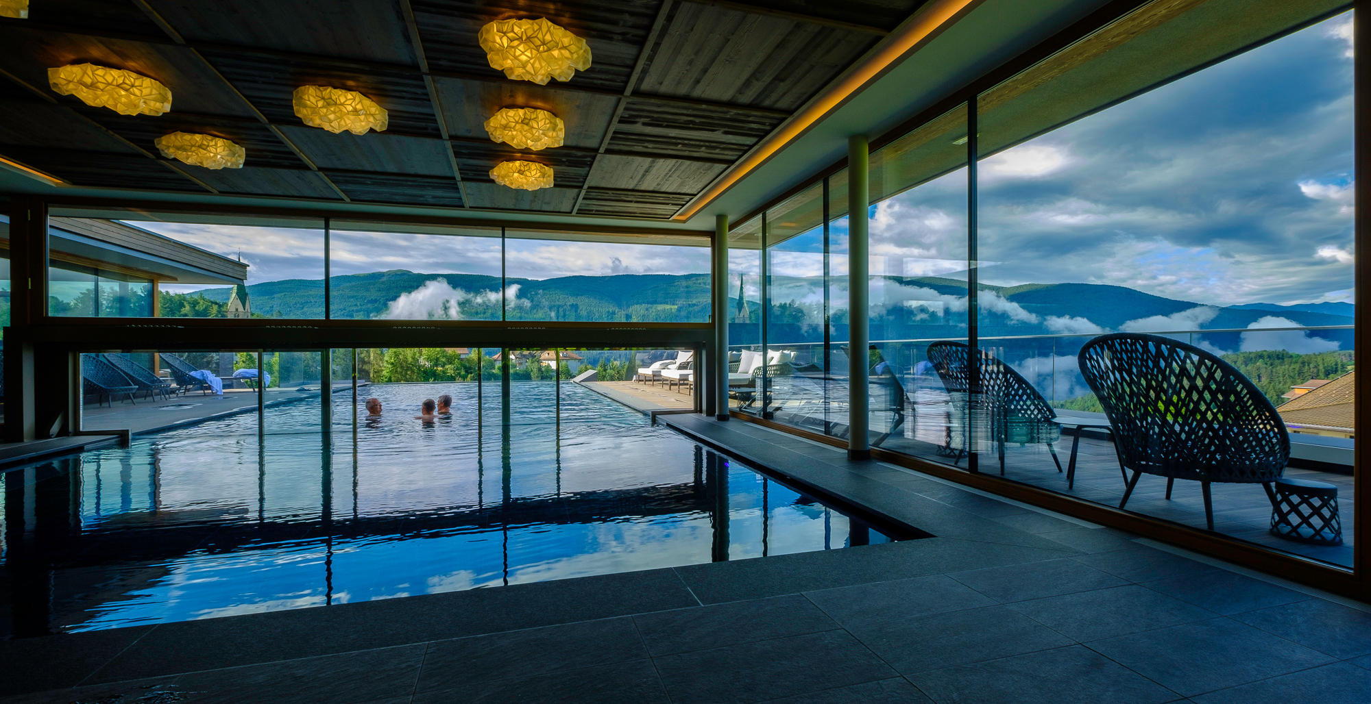 Indoor pool with panoramic view of the mountains and clouds at the Hotel Terentnerhof, with modern chairs and large windows.