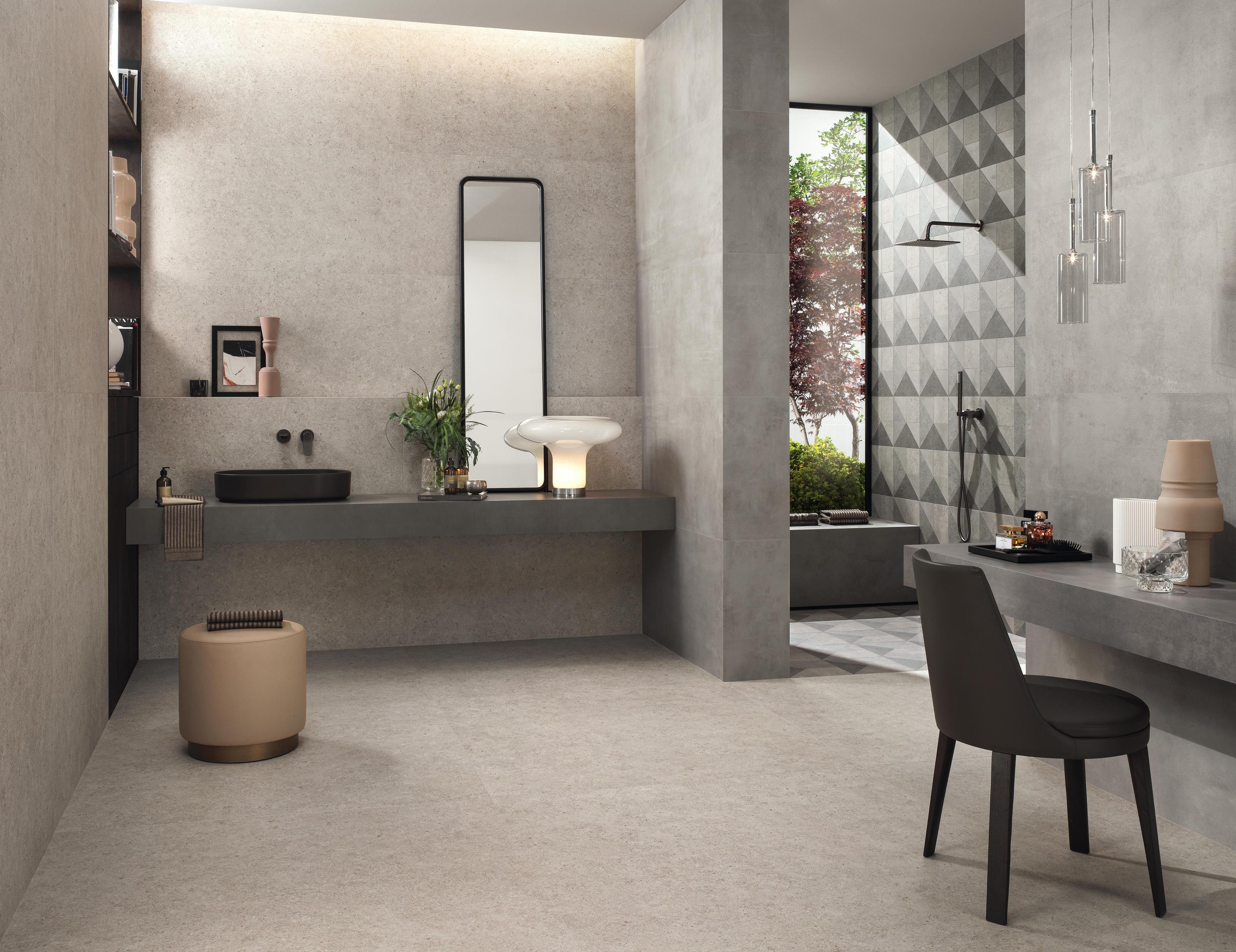 Salle de bain élégante avec carrelage gris effet pierre, lavabo noir, miroir vertical et douche avec carrelage à motifs géométriques.