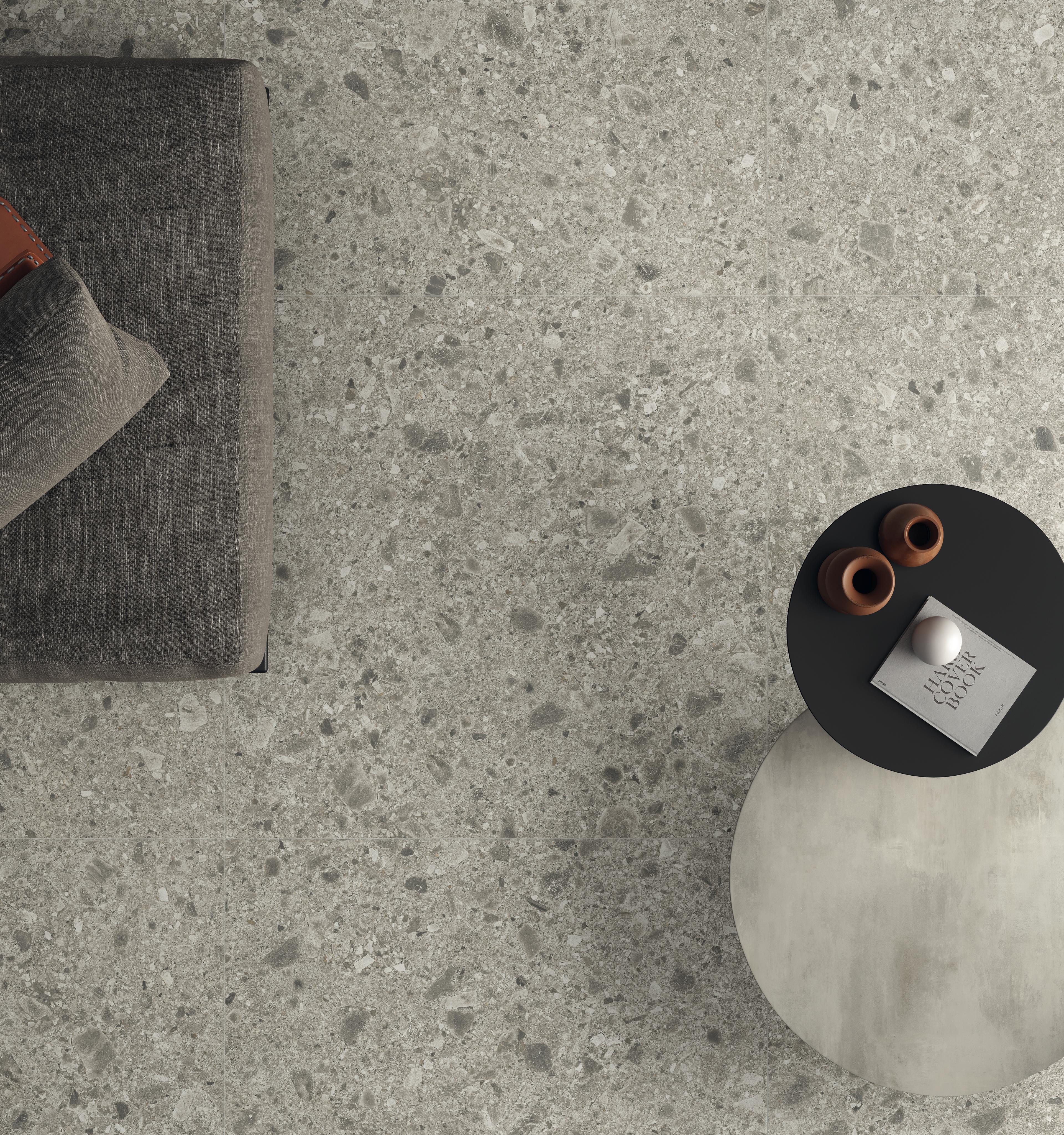 Detail des Bodens aus Gre-Stamm mit grauer Oberfläche und körniger Textur, eingerichtet mit einem dunkelgrauen Sofa und einem schwarzen runden Tisch mit dekorativen Accessoires.