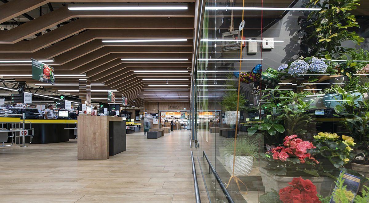 Интерьер супермаркета с секцией растений и цветов справа, полки продуктов и ящики внизу, с современным деревянным потолком.