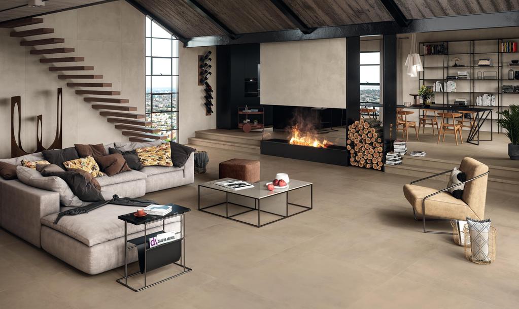 Moderna sala de estar con suelo de cerámica Atlas Concorde Boost Pro, escalera abierta de madera, amplio sofá gris con cojines decorativos y chimenea de leña. 
