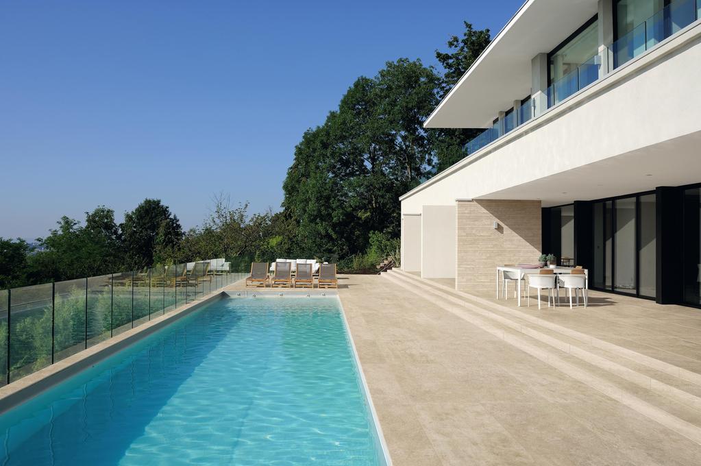 Piscina moderna circondata da piastrelle antiscivolo beige, area con lettini per prendere il sole e tavolo da pranzo bianco. 