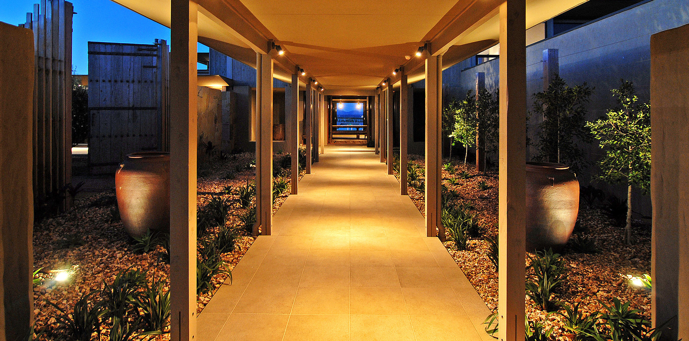Couloir extérieur éclairé d’une villa privée en Australie, avec sol en pierre, colonnes de soutien et jardin avec plantes décoratives.