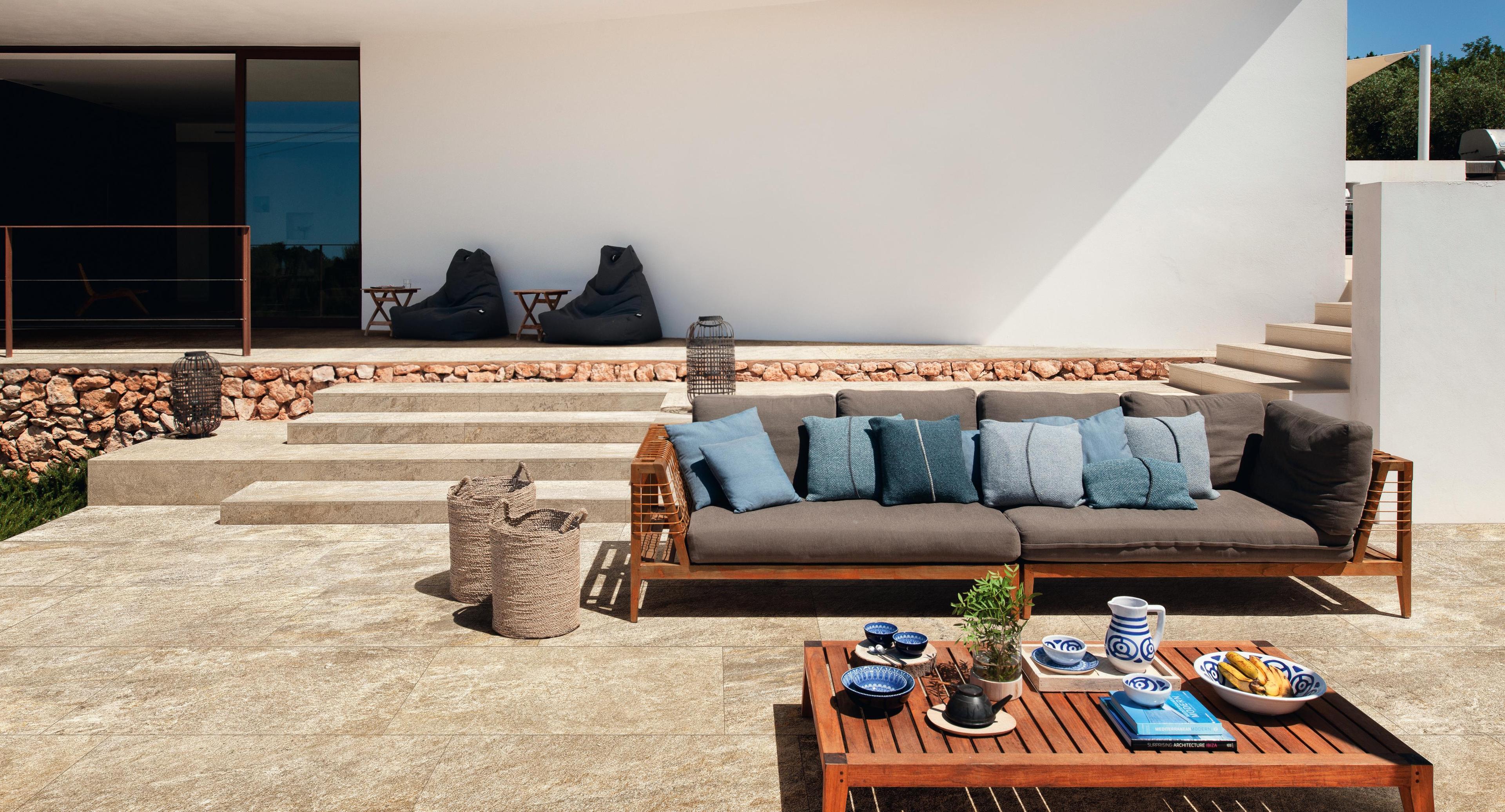  Floor of an outdoor terrace covered with yellow tiles in porcelain stoneware effect, sofa with blue and gray cushions, and table with decorations.